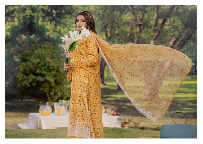 A woman in a yellow floral dress stands outdoors holding a bouquet of white lilies. A flowy scarf extends from her dress, and picnic items are visible in the background.